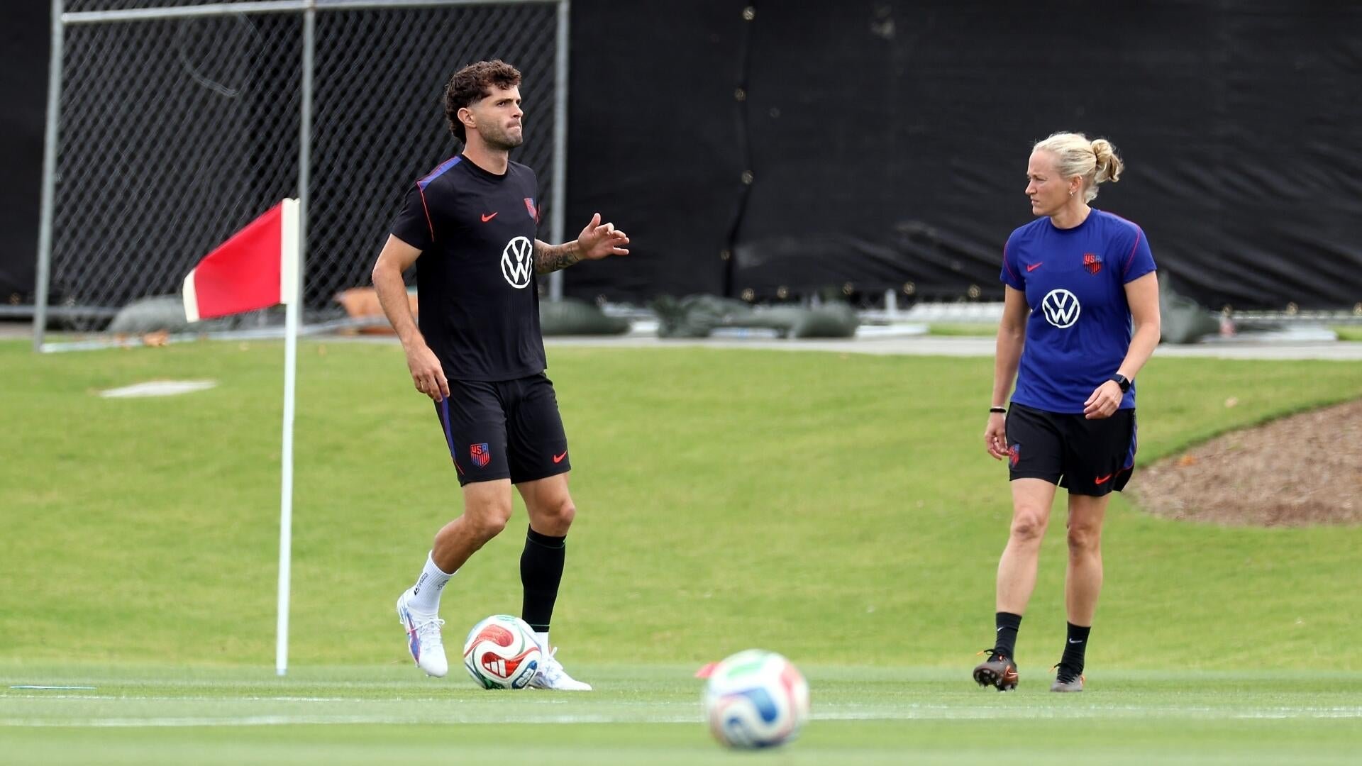 A man and a woman in USA soccer training gear stand on a grass field, with the man gesturing near a corner flag.