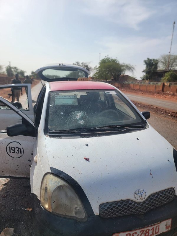 A white Toyota taxi with license plate "BS:7810" and a shattered windshield sits parked on a roadside with its driver-side door open.