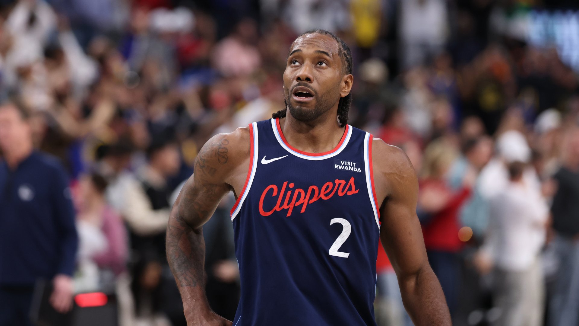 A basketball player wearing a dark blue Clippers jersey with the number 2 looks upward during a game, with a blurred crowd in the background.