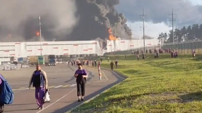People walk away from a large industrial building engulfed in flames and thick black smoke.