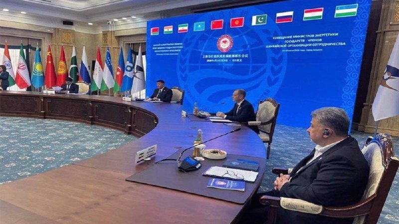 Officials sit at a large oval conference table in a formal meeting room, with national flags and a blue backdrop displaying SCO summit branding.