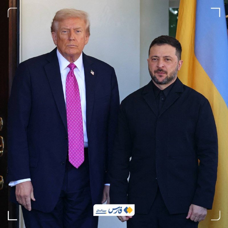 Two men in dark suits stand side by side, one wearing a pink tie, with a blue and yellow flag visible behind them.