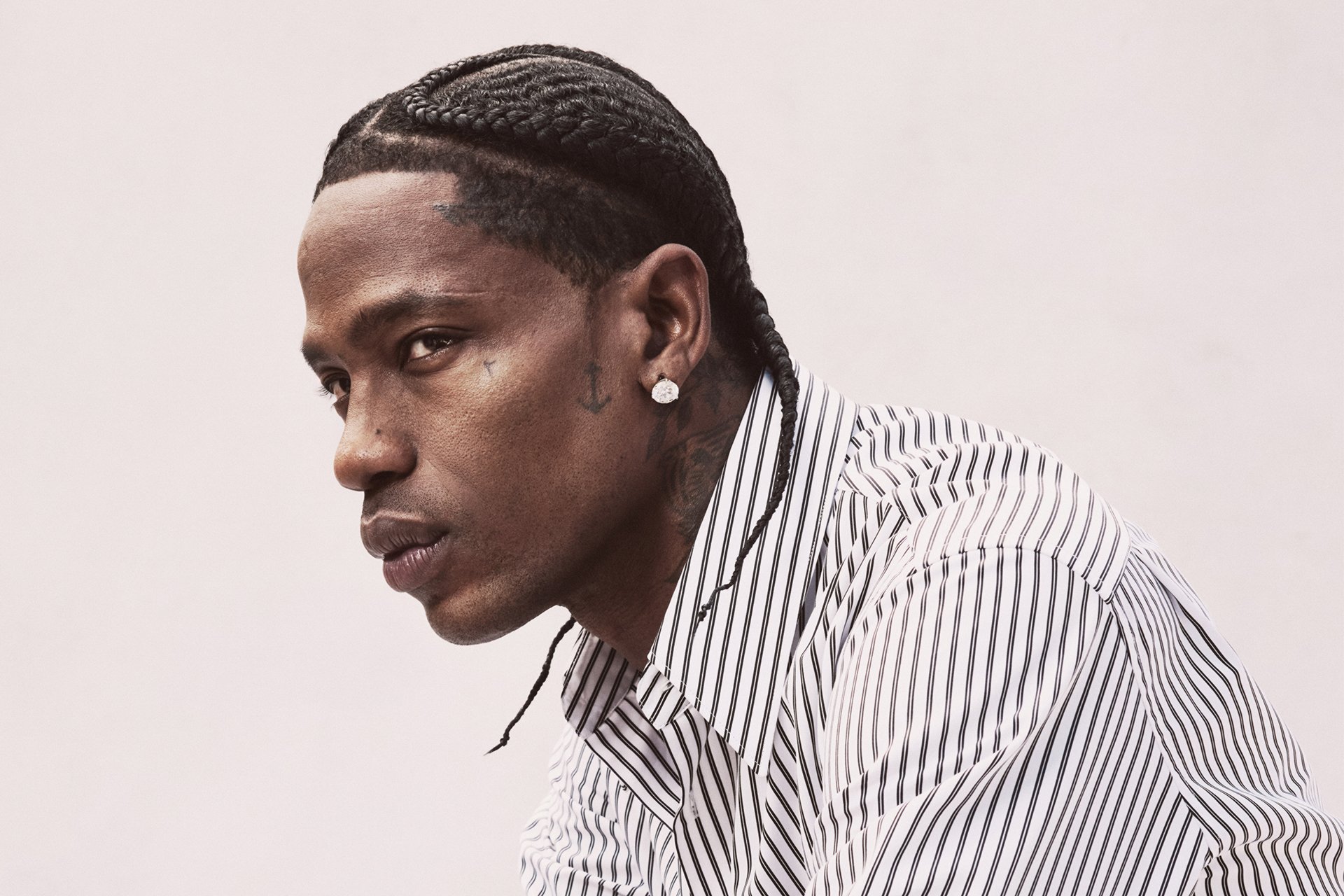 A man with braided hair, facial and neck tattoos, a diamond earring, and a white striped shirt poses against a plain light background.