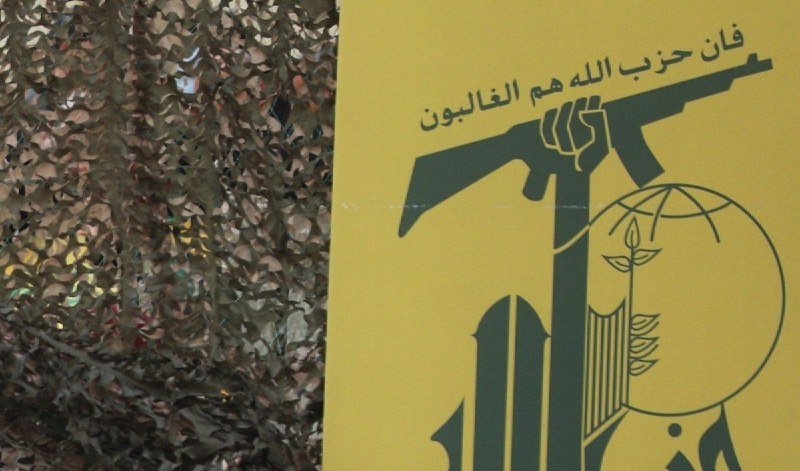 A yellow banner with Arabic script and a green graphic depicting a fist gripping a rifle, missile, and globe hangs beside camouflage netting.
