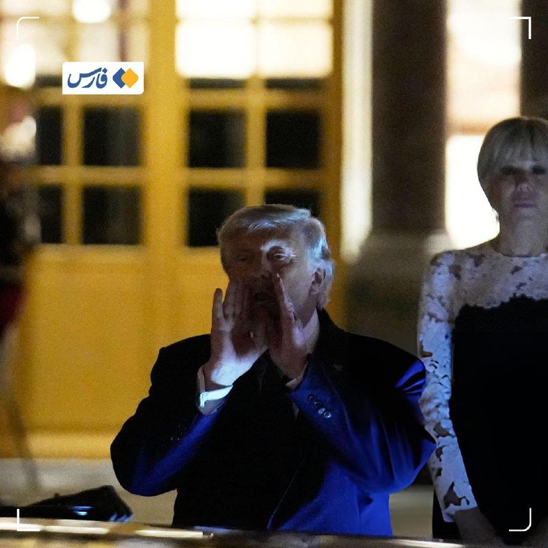 A white-haired man in a dark suit holds his hands near his face beside a blonde woman in a black and white dress, illuminated in a dim setting.