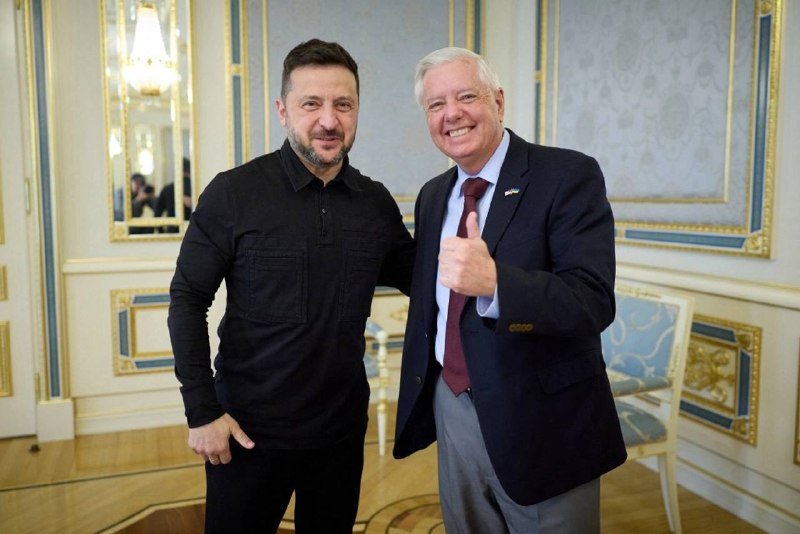 Two men pose smiling in an ornately decorated room with gold-trimmed blue walls and a mirror, one in a black shirt and the other in a dark suit giving a thumbs-up.