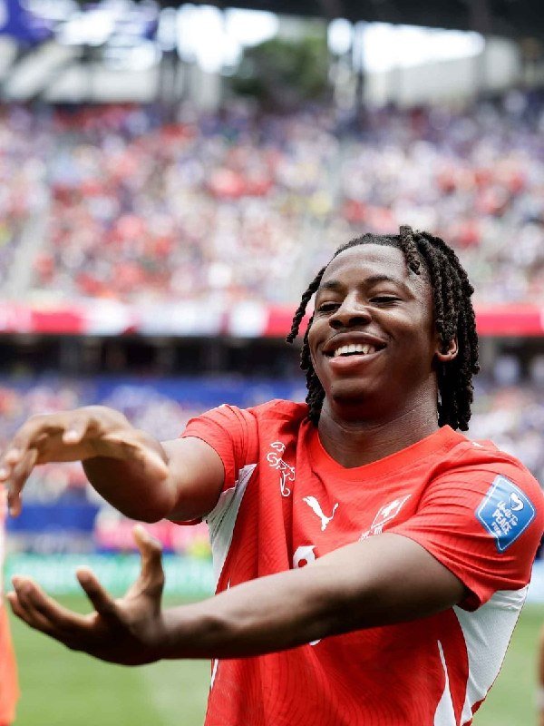 A smiling soccer player with braided hair wearing a red and white jersey extends his arms outward in celebration on the field before a stadium crowd.