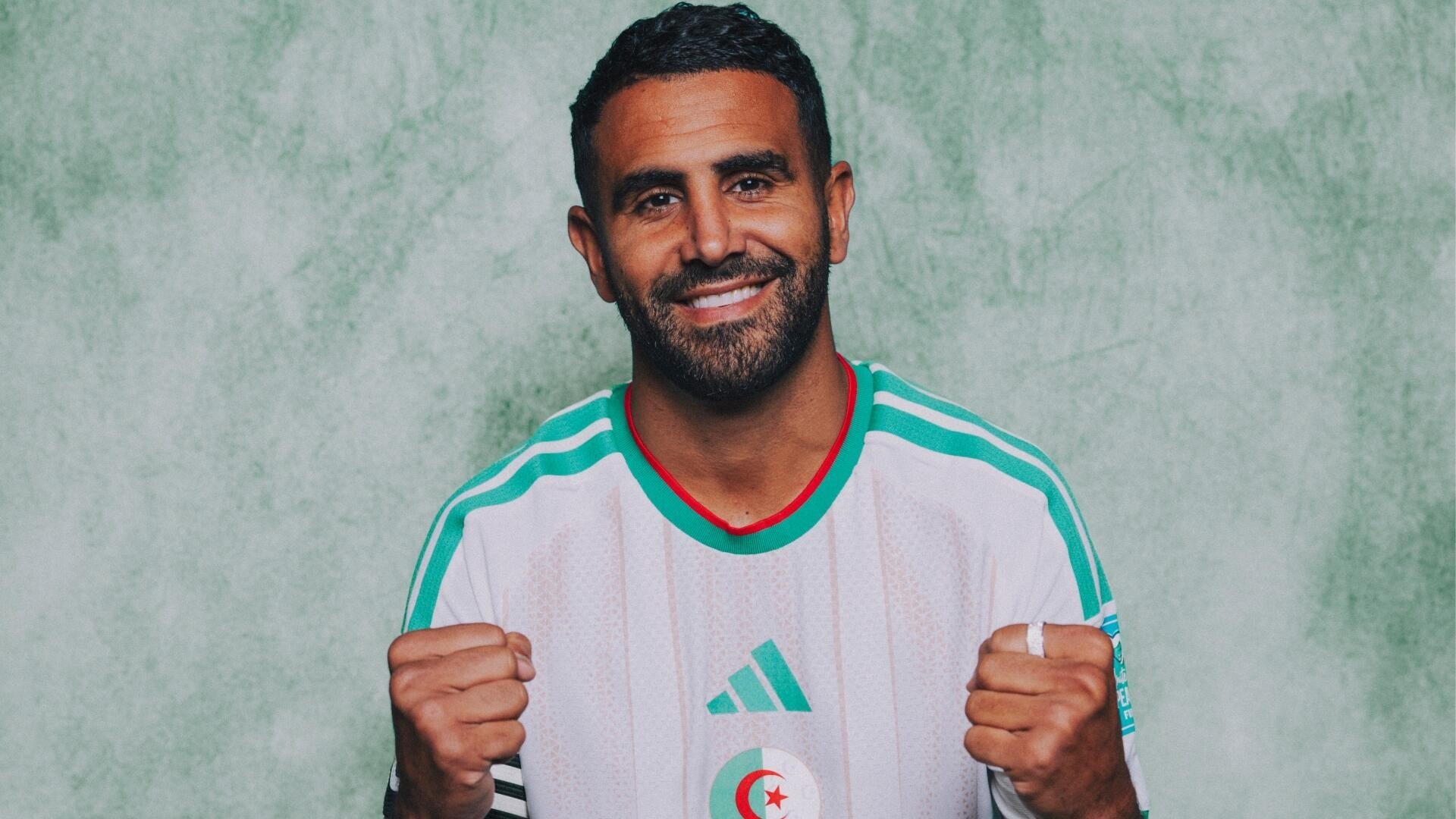 A smiling, bearded man wearing a white and green Adidas jersey with a crescent and star emblem poses with clenched fists against a green background.
