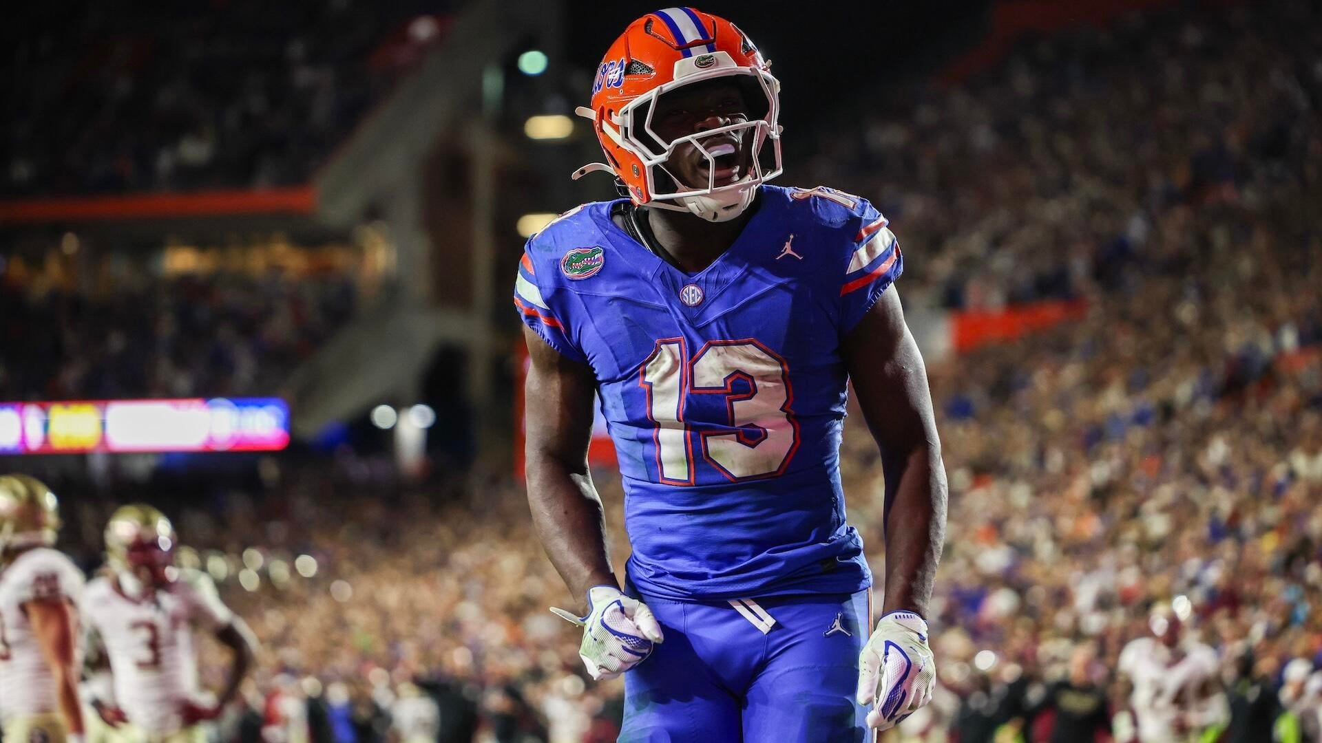 A football player in a blue #13 jersey and orange helmet yells with his mouth wide open during a nighttime game in a crowded stadium.