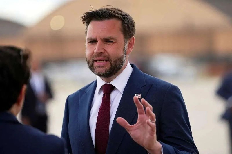 A bearded man in a blue suit, white shirt, and burgundy tie with an American flag pin gestures while speaking outdoors near a blurred building.