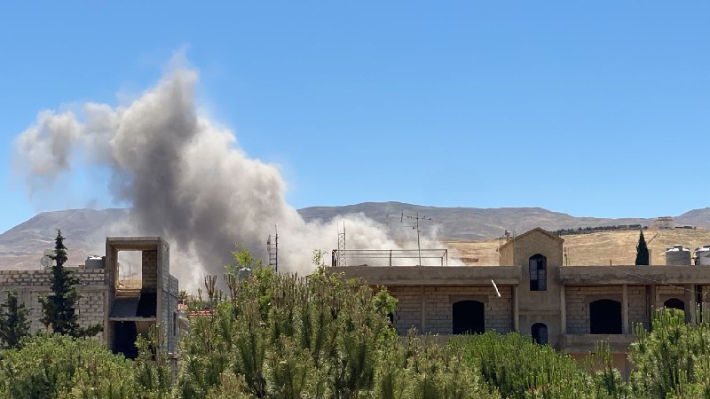 A large gray plume of smoke rises above a densely built hillside town with white and beige buildings under a clear blue sky, with a tree in the foreground.
