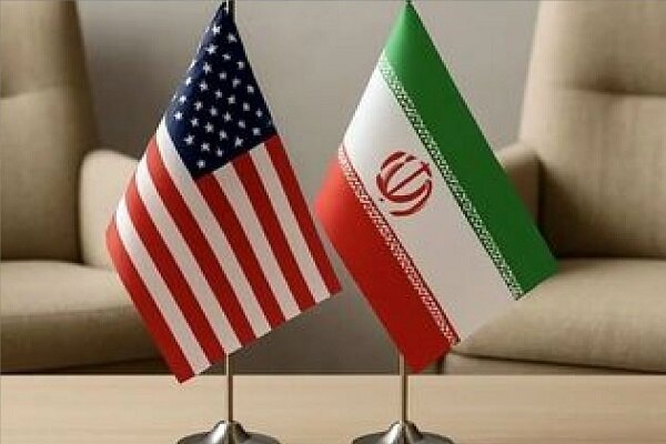 Small desktop flags of the United States and Iran stand side by side on a light-colored table in front of beige upholstered chairs.