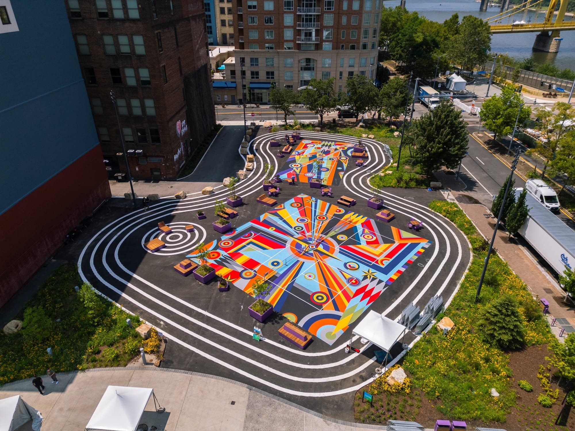 An aerial view shows a public plaza featuring large, colorful geometric murals surrounded by concentric white painted lines, with purple benches, nearby brick buildings, and a yellow suspension bridge in the background.