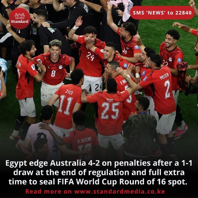 A group of soccer players in red jerseys huddle together on a grass field, raising their arms and embracing in celebration.