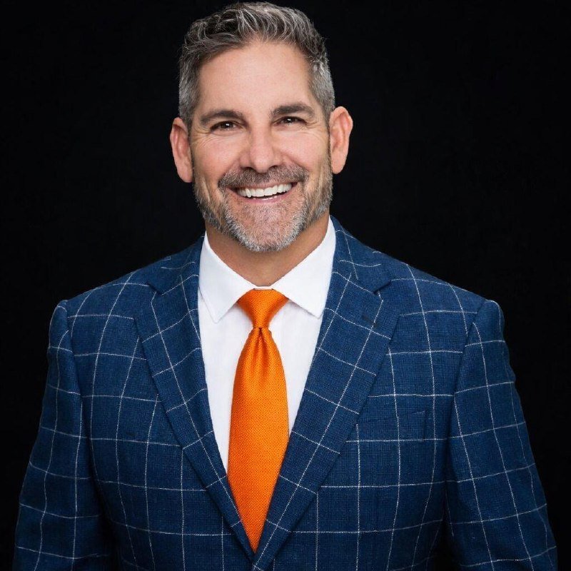 A smiling man with a graying beard wears a blue windowpane-checked suit, white shirt, and bright orange tie against a black background.