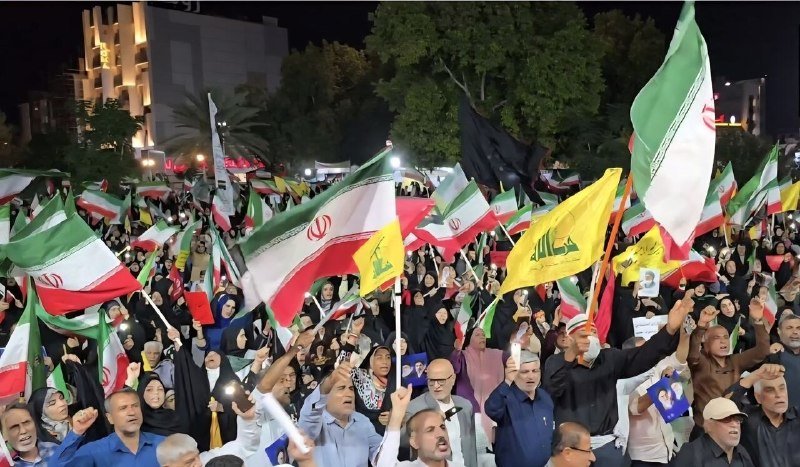 A bespectacled man in a gray suit speaks into a microphone, with a yellow and green flag and Arabic-script banner visible in the background.