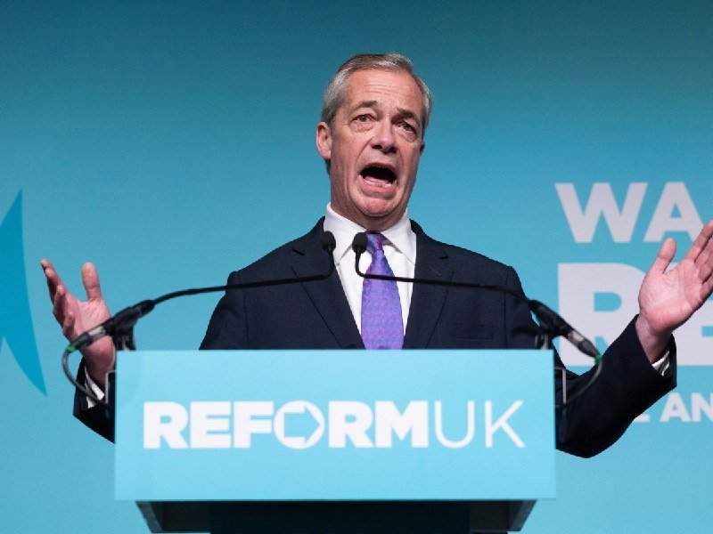 A gray-haired man in a dark suit and patterned tie speaks animatedly at a podium labeled "REFORMUK," gesturing with both hands raised.