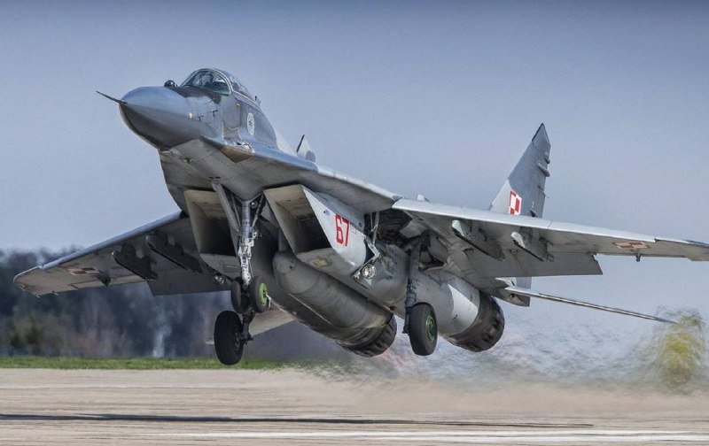 A gray military fighter jet with swept wings and deployed landing gear lifts off from a runway, kicking up dust.