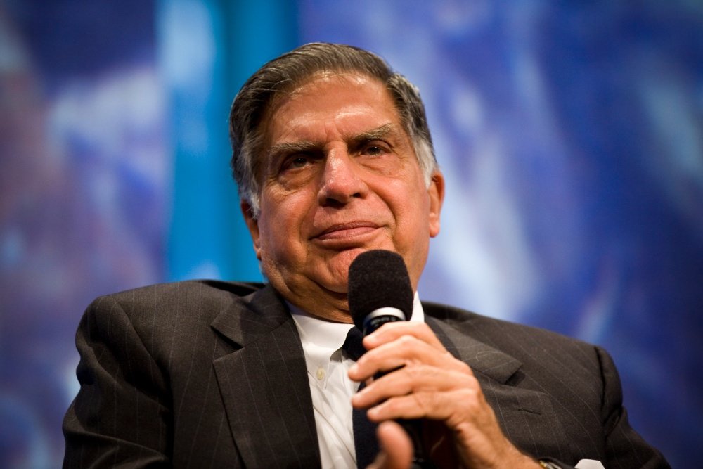 An elderly man in a dark suit holds a microphone near his face against a blurred blue background, appearing to speak at an event.