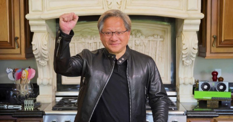 A man in a black leather jacket raises his fist while smiling in a kitchen featuring an ornate stone fireplace and cookware displayed on the counter.