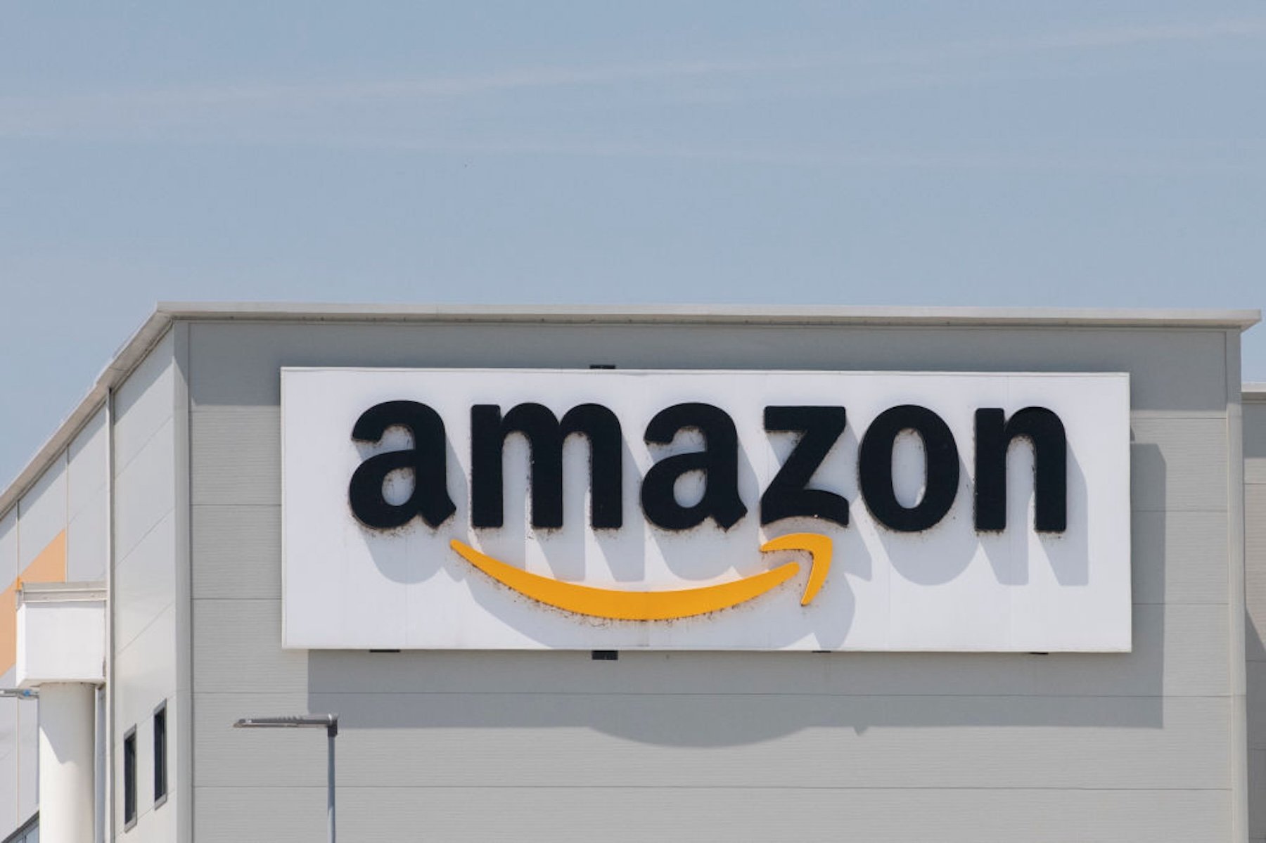A large Amazon logo sign is mounted on the exterior wall of a gray industrial building under a clear blue sky.
