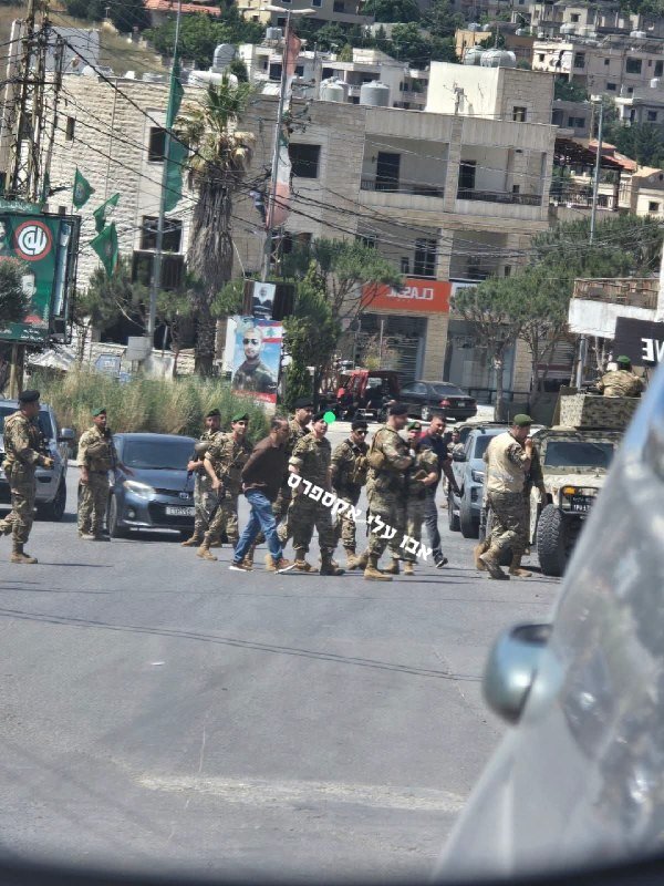 Armed soldiers in camouflage uniforms escort a handcuffed civilian in jeans across a paved street, with military vehicles, parked cars, and hillside buildings visible in the background.