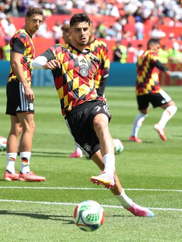 Soccer players in red, yellow, and black checkered jerseys warm up on a grass field, with one kicking a ball while spectators watch from red stadium seats in the background.