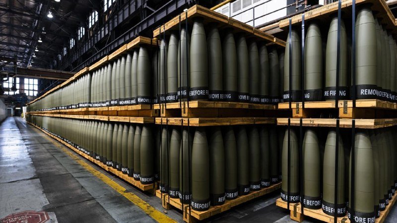 Rows of olive-green artillery shells labeled "REMOVE BEFORE FIRING" are stacked on wooden pallets inside an industrial warehouse.