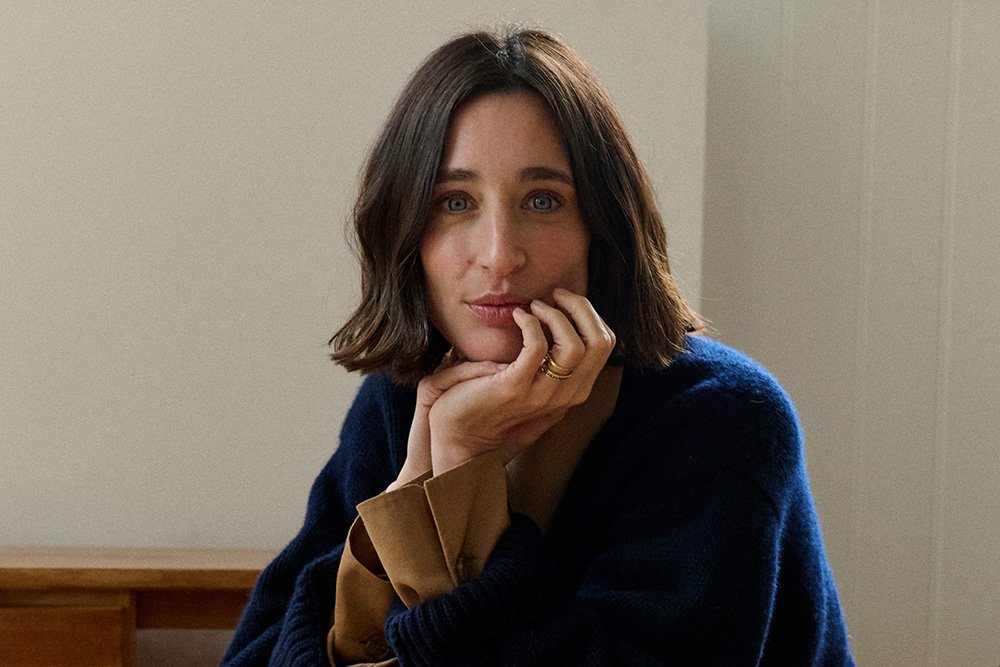 A woman with shoulder-length dark hair rests her chin on her hand, wearing a navy cardigan over a tan top against a neutral background.