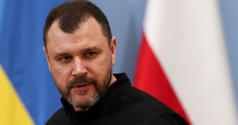 A bearded man in a black jacket is shown in close-up with blue, yellow, and red-and-white flags visible in the background.