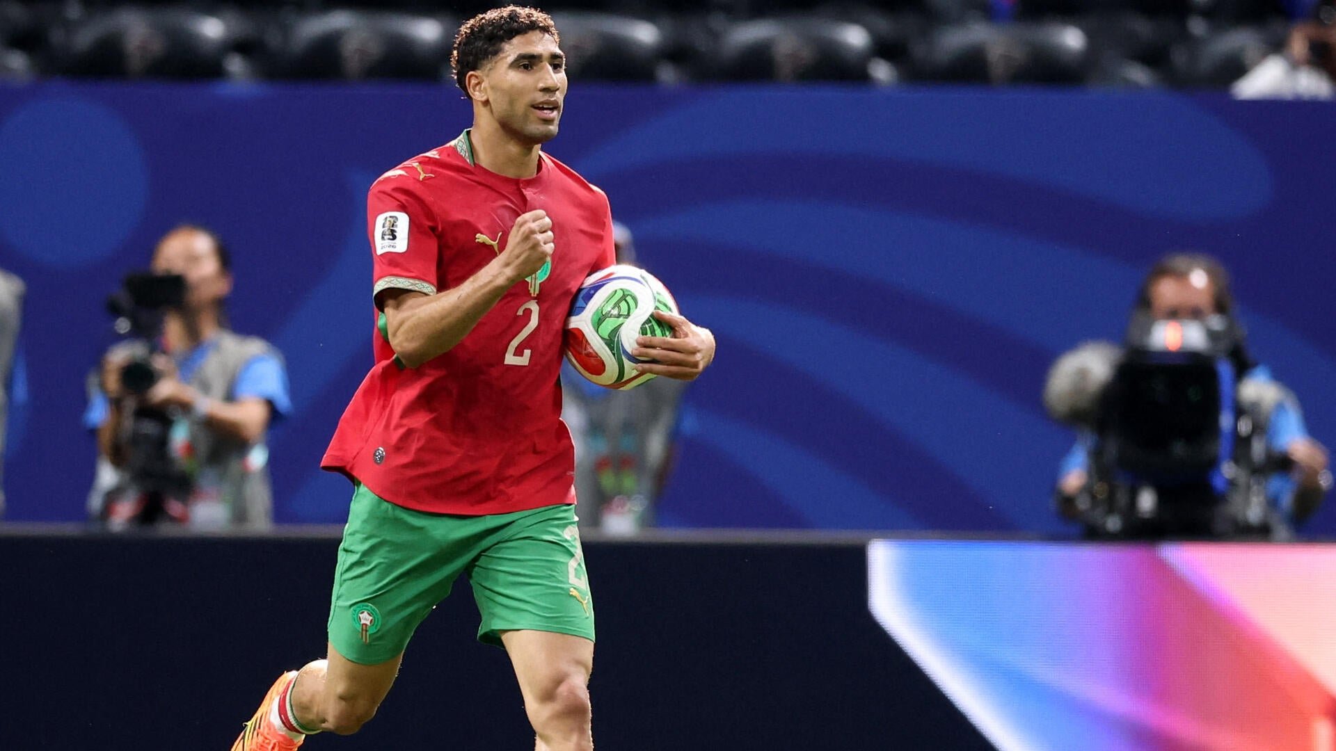 A soccer player wearing a red Morocco jersey with the number 2 runs while holding a colorful match ball, with camera operators visible in the background.