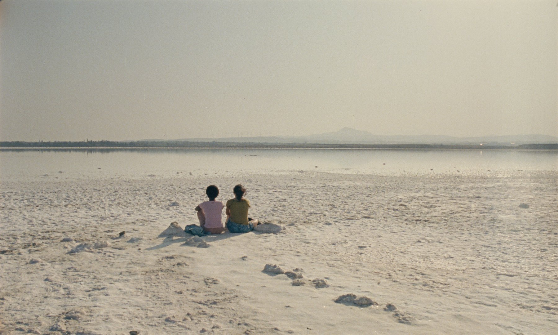 Two people sit side by side on a white, crystalline shore facing a calm body of water with distant mountains visible on the horizon.