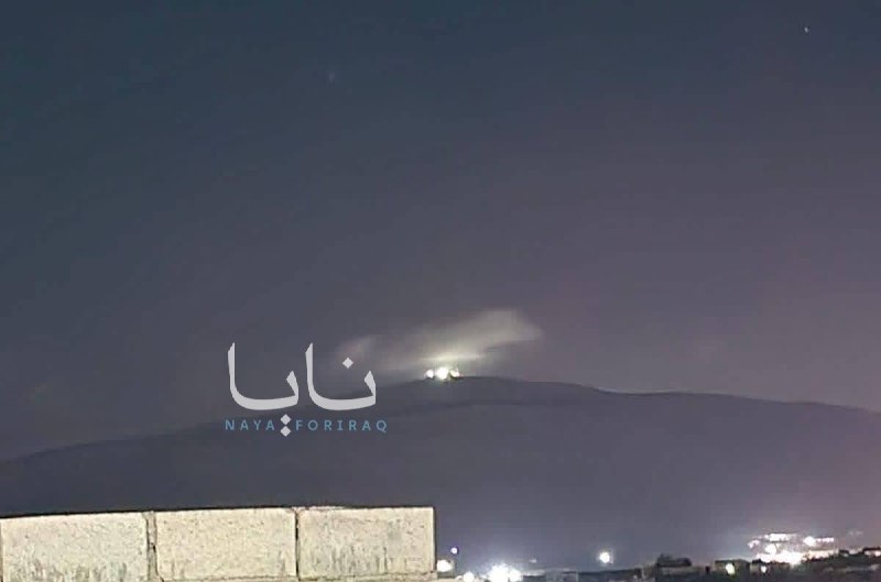 The Iranian national flag waves against a blurred cityscape background, displaying its green, white, and red horizontal stripes with the red emblem in the center.