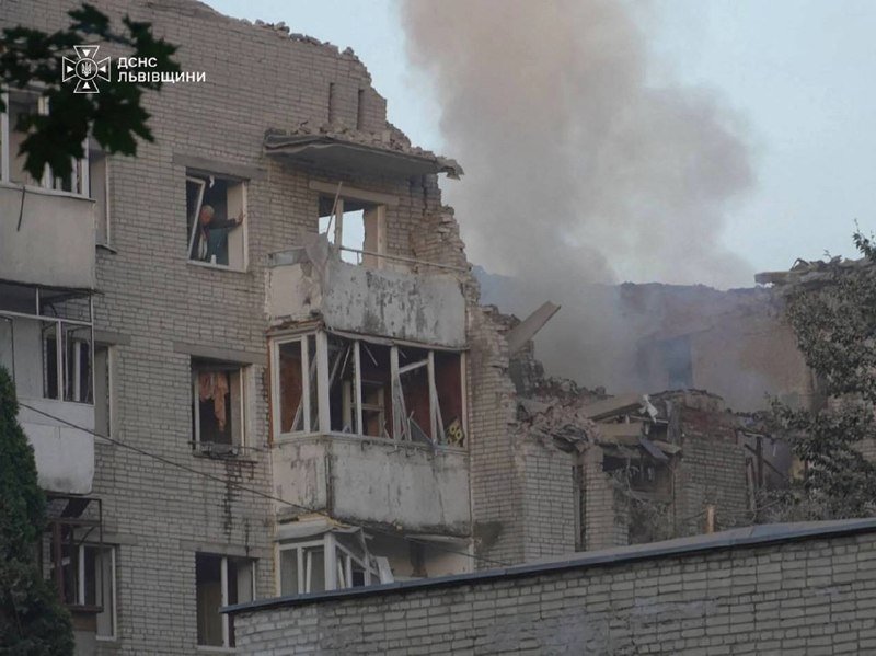 A heavily damaged multi-story brick apartment building stands with shattered windows, exposed interiors, and crumbling walls, while thick smoke rises from the structure behind it.