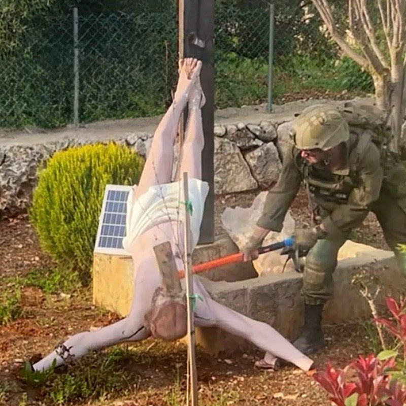 A soldier in green uniform and helmet uses a long orange tool on a white figure suspended upside-down from a wooden post in a garden setting.