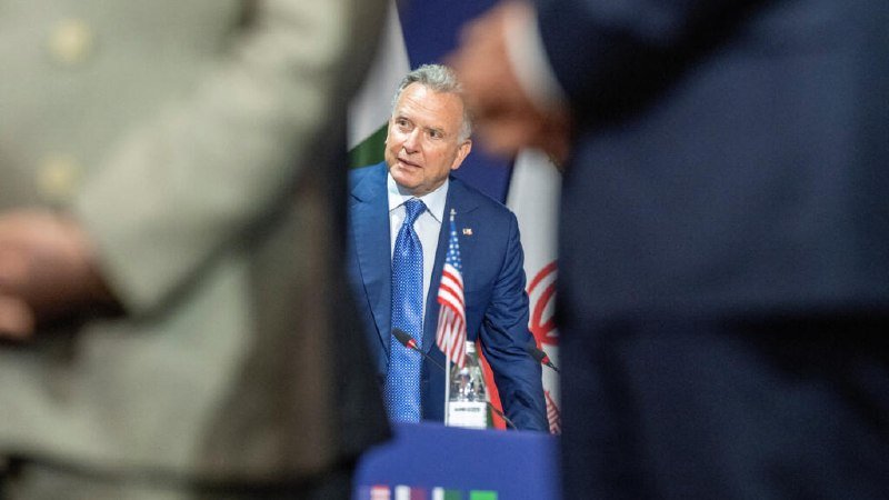A man in a blue suit and tie stands at a podium with a small American flag, viewed between two blurred figures in the foreground.