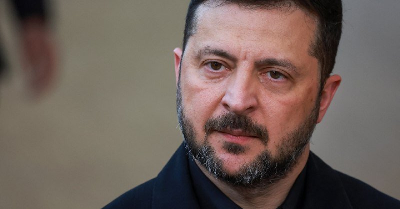 A close-up shows a man with a dark beard and short hair wearing a dark coat, looking off to the side against a blurred background with another partially visible figure.