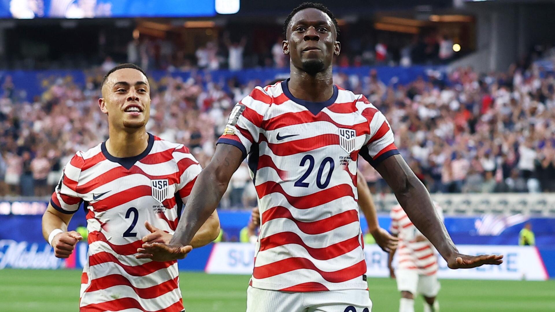 A soccer player in a dark USA jersey with a FIFA World Cup 2026 patch applauds with a serious expression against a blurred stadium background.