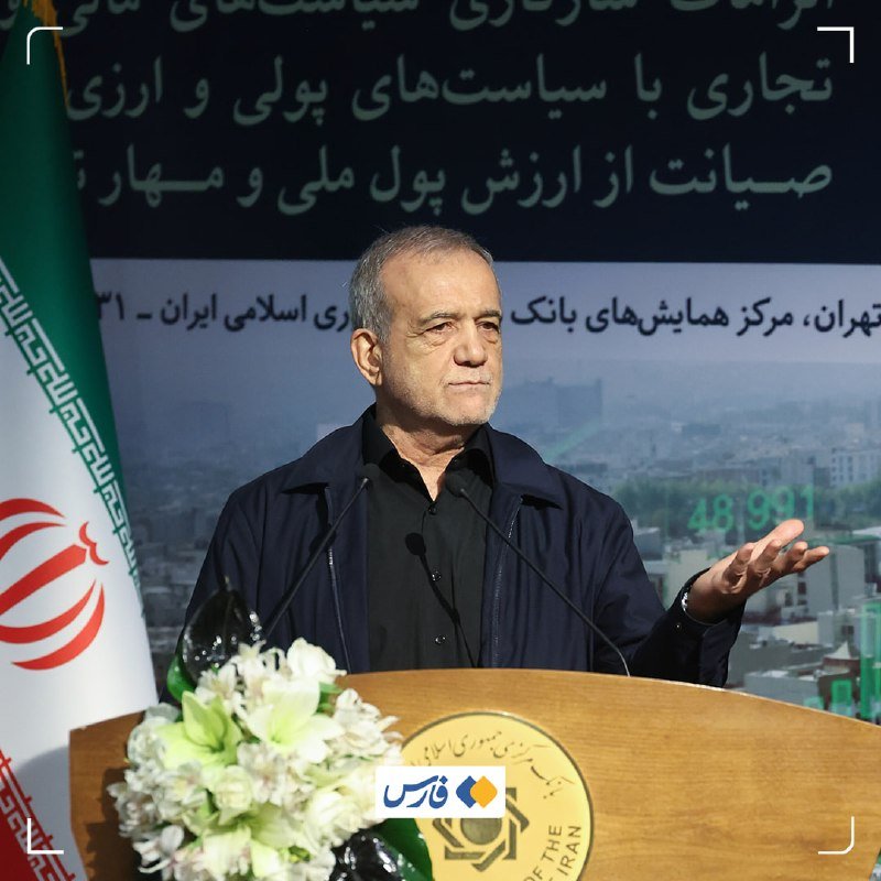 A gray-haired man in a dark jacket gestures while speaking at a wooden podium with an Iranian flag and a Persian-text banner behind him.