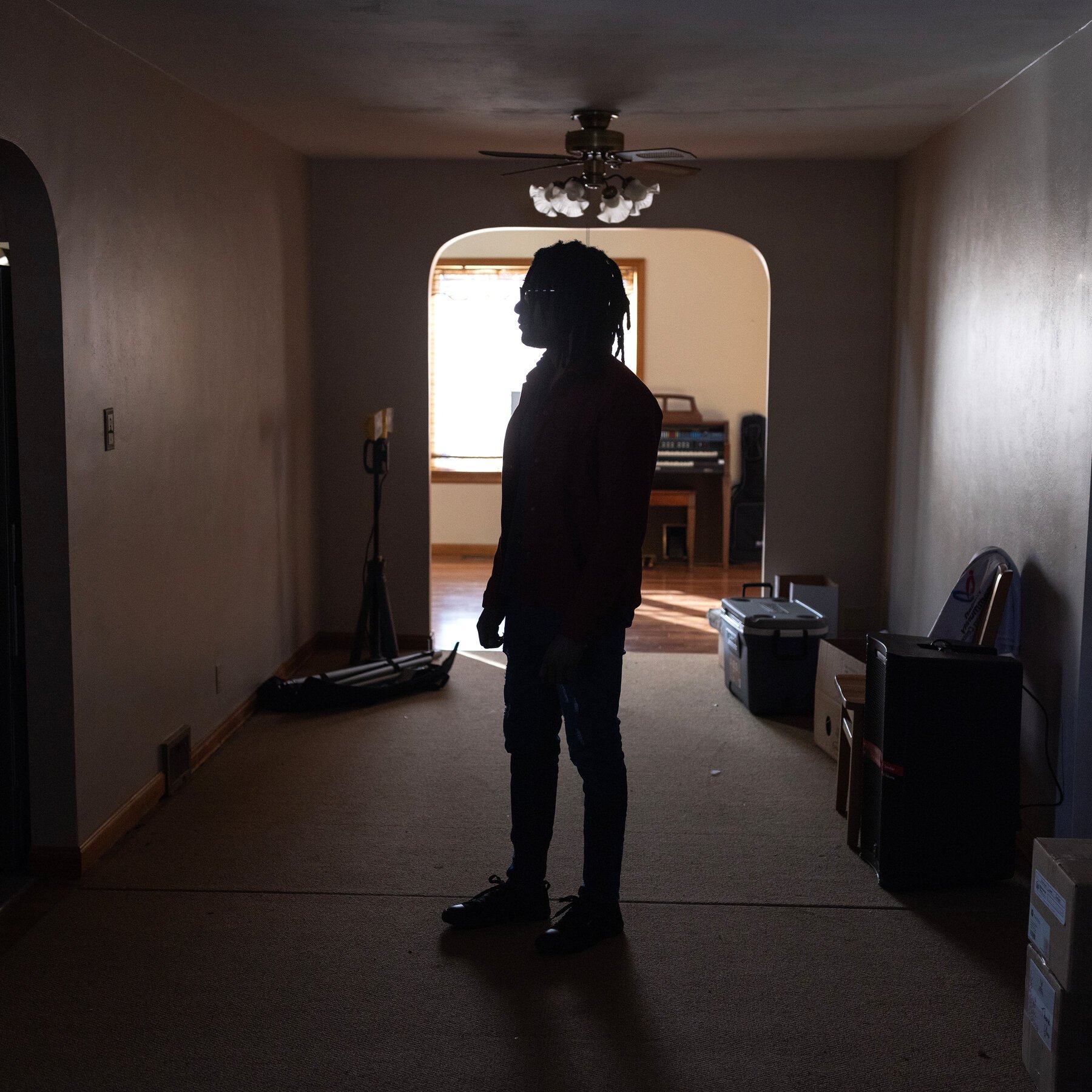 A silhouetted figure with long hair and glasses stands in a dimly lit hallway, facing an archway that leads to a sunlit room containing a keyboard instrument, with stacked boxes and equipment along the walls.