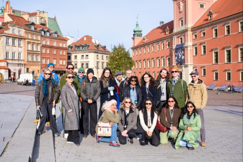 A group of approximately eighteen people pose together in a cobblestone plaza surrounded by colorful historic buildings with red-tiled roofs and a green-domed tower.