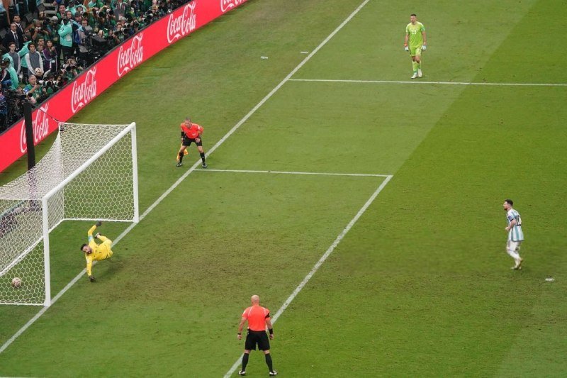A soccer goalkeeper dives toward the ball near the goal line while players in striped, green, and orange jerseys watch during a match in front of advertising boards.