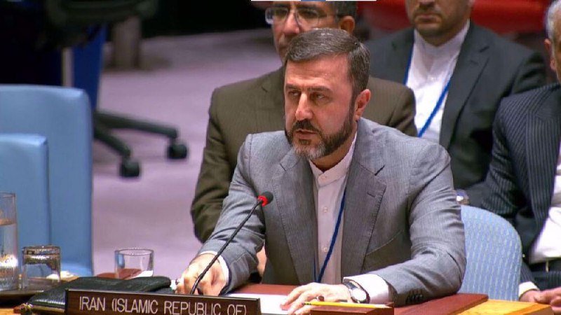 A bearded man in a gray suit speaks into a microphone at a desk labeled "IRAN (ISLAMIC REPUBLIC OF)," with other officials seated behind him.