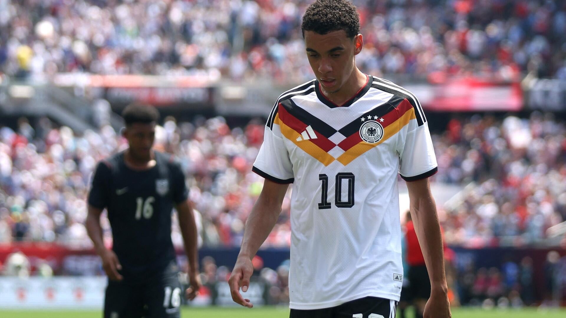 A smiling soccer player wearing an orange Puma jersey with the number 11 and a FIFA World Cup 2026 patch clenches both fists in celebration before a stadium crowd.