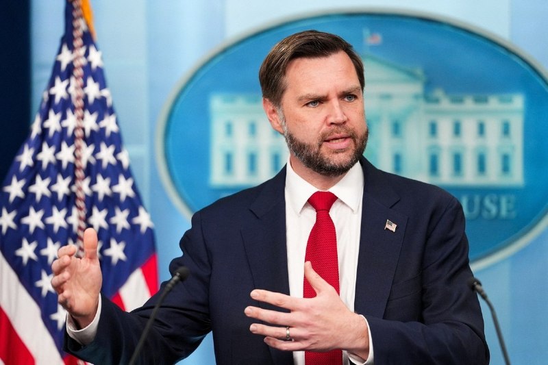 A gray-bearded man in glasses, a navy blazer, and blue shirt gestures with his hands while speaking in an office setting.