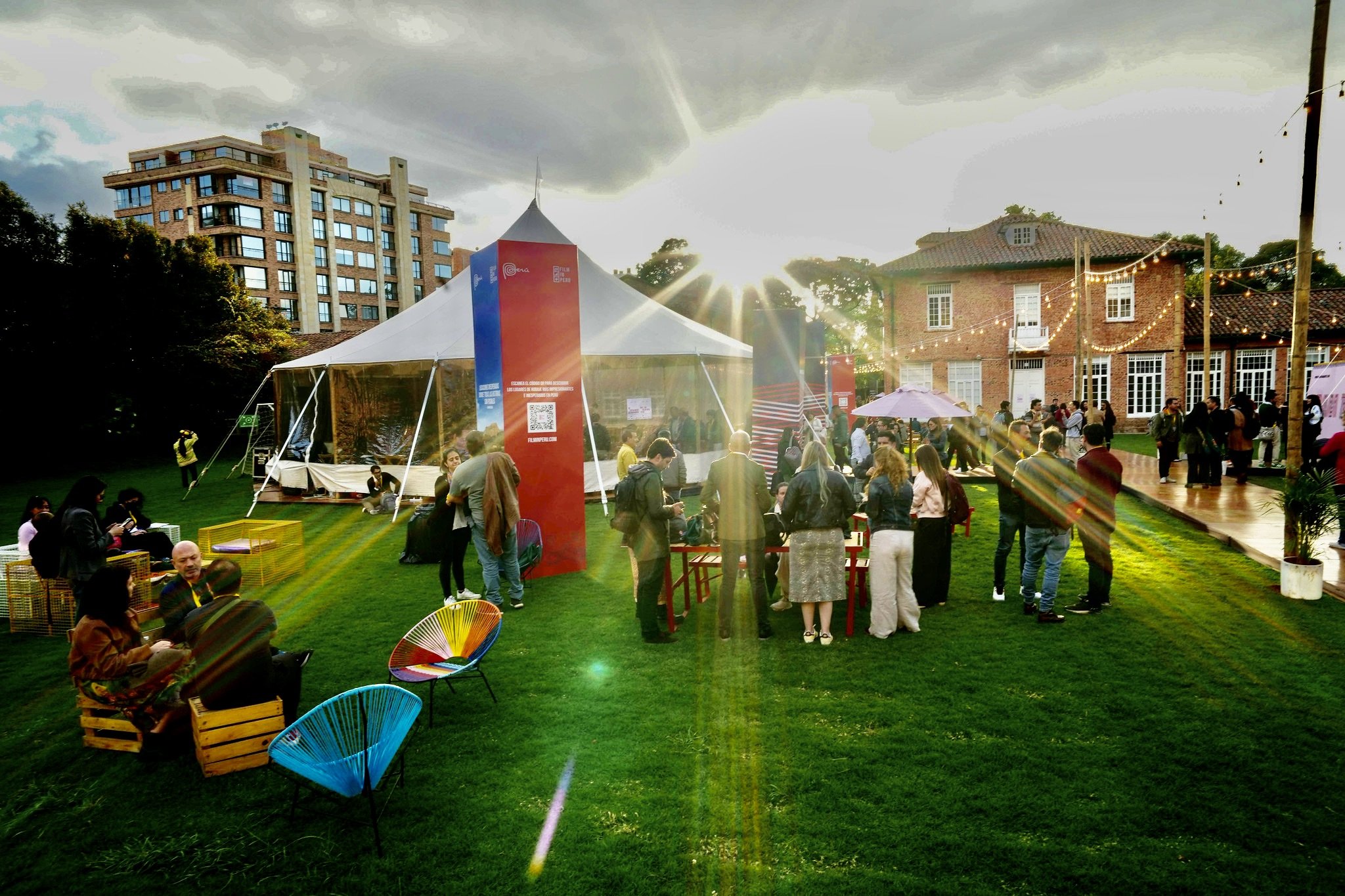 People gather on a grassy lawn at an outdoor film festival event under a large white tent, with string lights, a brick building, and apartment tower in the background as the sun sets.