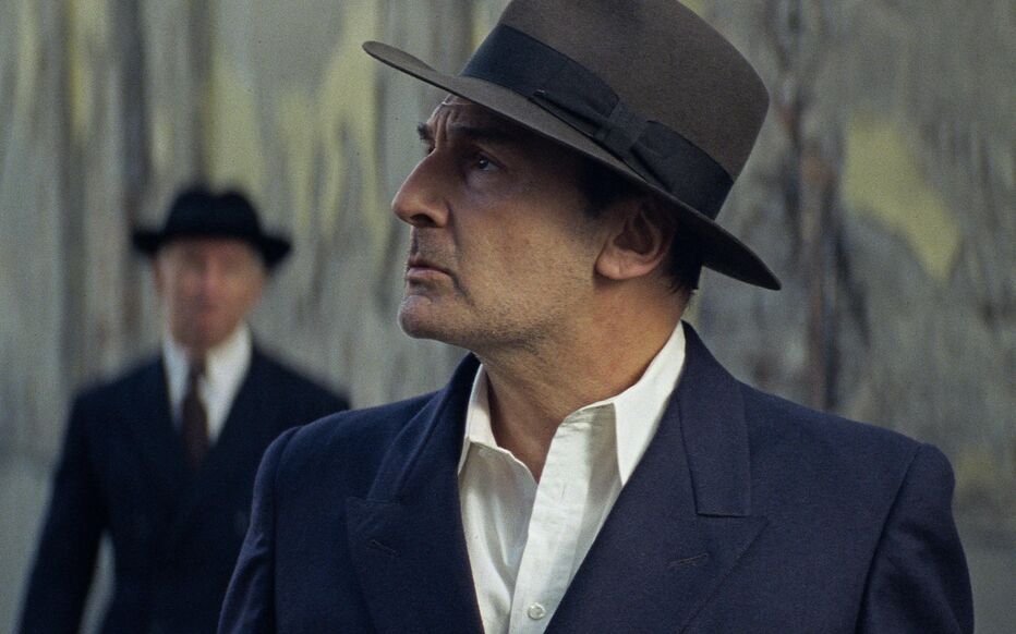 A man in a fedora and dark suit stands in the foreground while another man in a hat and suit appears blurred behind him.