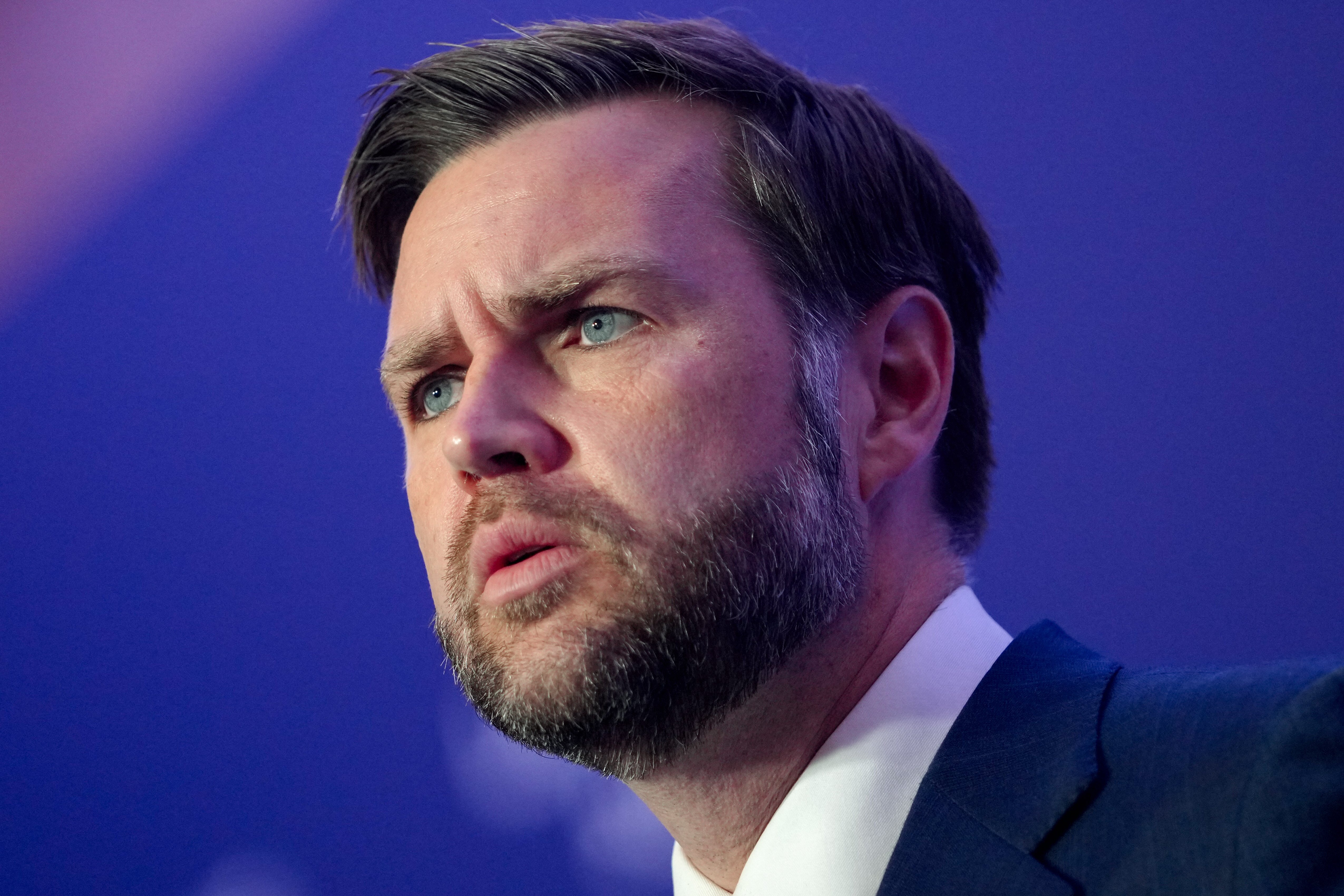 A bearded man in a dark blue suit and white shirt speaks against a blurred purple background.