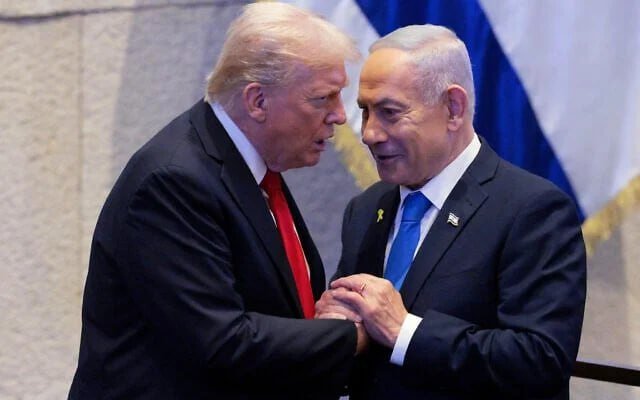 Two men in suits—one with a red tie and blonde hair, the other with gray hair and a blue tie—shake hands and smile in front of an Israeli flag.