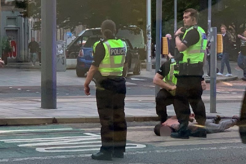 Three police officers in high-visibility vests attend to a person lying on a city street, with bystanders and parked vehicles visible in the background.